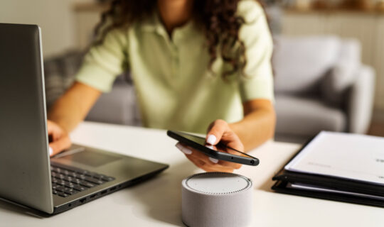 A woman in a light green short sleeve shirt uses a laptop computer, mobile phone, and an Alexa device.